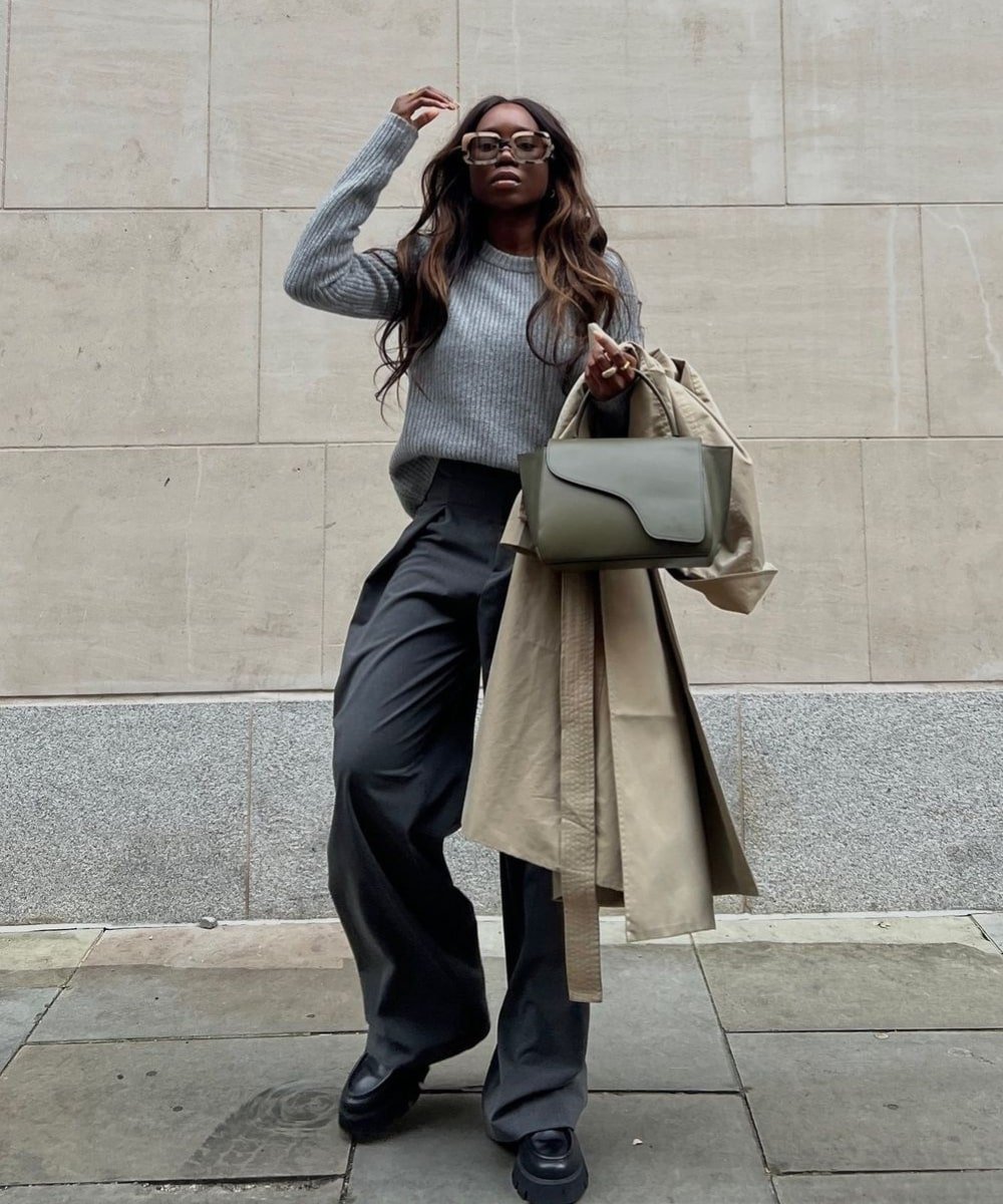 Danielle Jinadu - calça alfaiataria cinza, trico e óculos de sol - looks minimalistas - Inverno  - em pé na rua com um trench coat no braço - https://stealthelook.com.br