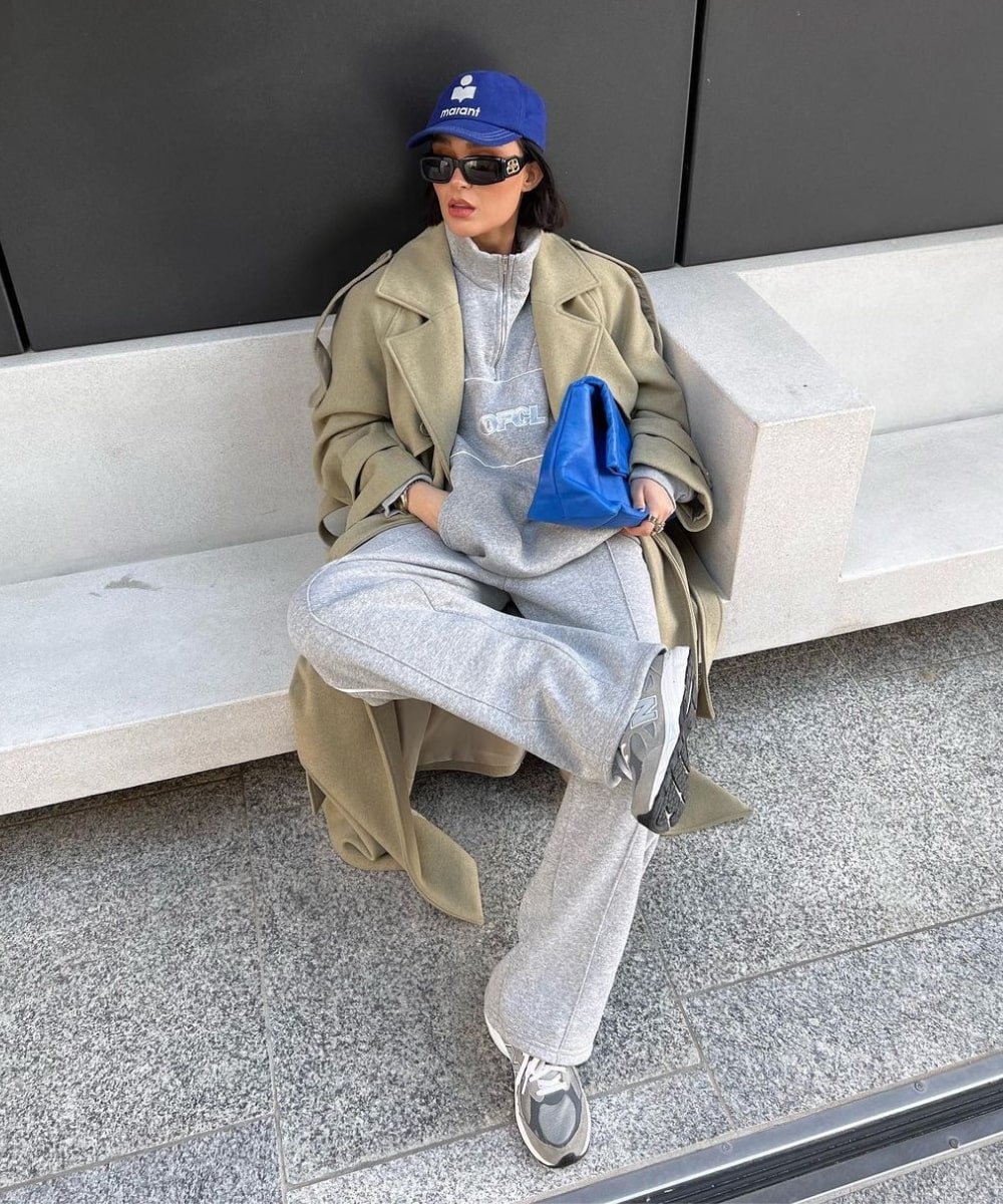 Alicia Roddy - conjunto de moletom cinza, trench coat, boné azul e óculos preto - looks com moletom - Inverno  - sentada em uma cadeira - https://stealthelook.com.br