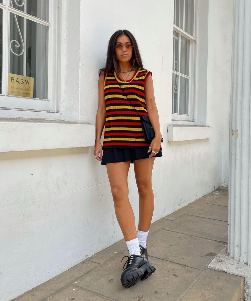 Marj Moore - saia curta preta, colete colorido de tricô e oxfords - sapato oxford - Verão - em pé na rua usando óculos de sol - https://stealthelook.com.br