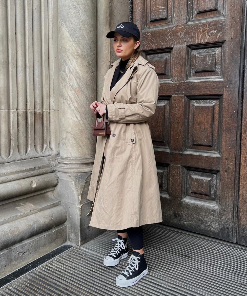 Isabella Duder - calça legging preta, blusa preta, trench coat e tênis - peças de alfaiataria - Inverno  - em pé na rua usando um boné - https://stealthelook.com.br