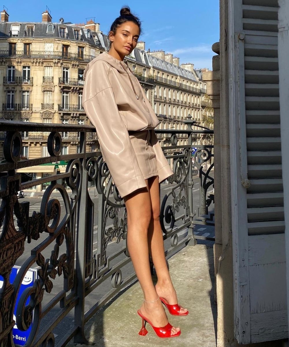 Amina Muaddi - vestido curto bege de manga longa e sapato vermelho - sapato tendência - Verão - em pé em uma varanda - https://stealthelook.com.br