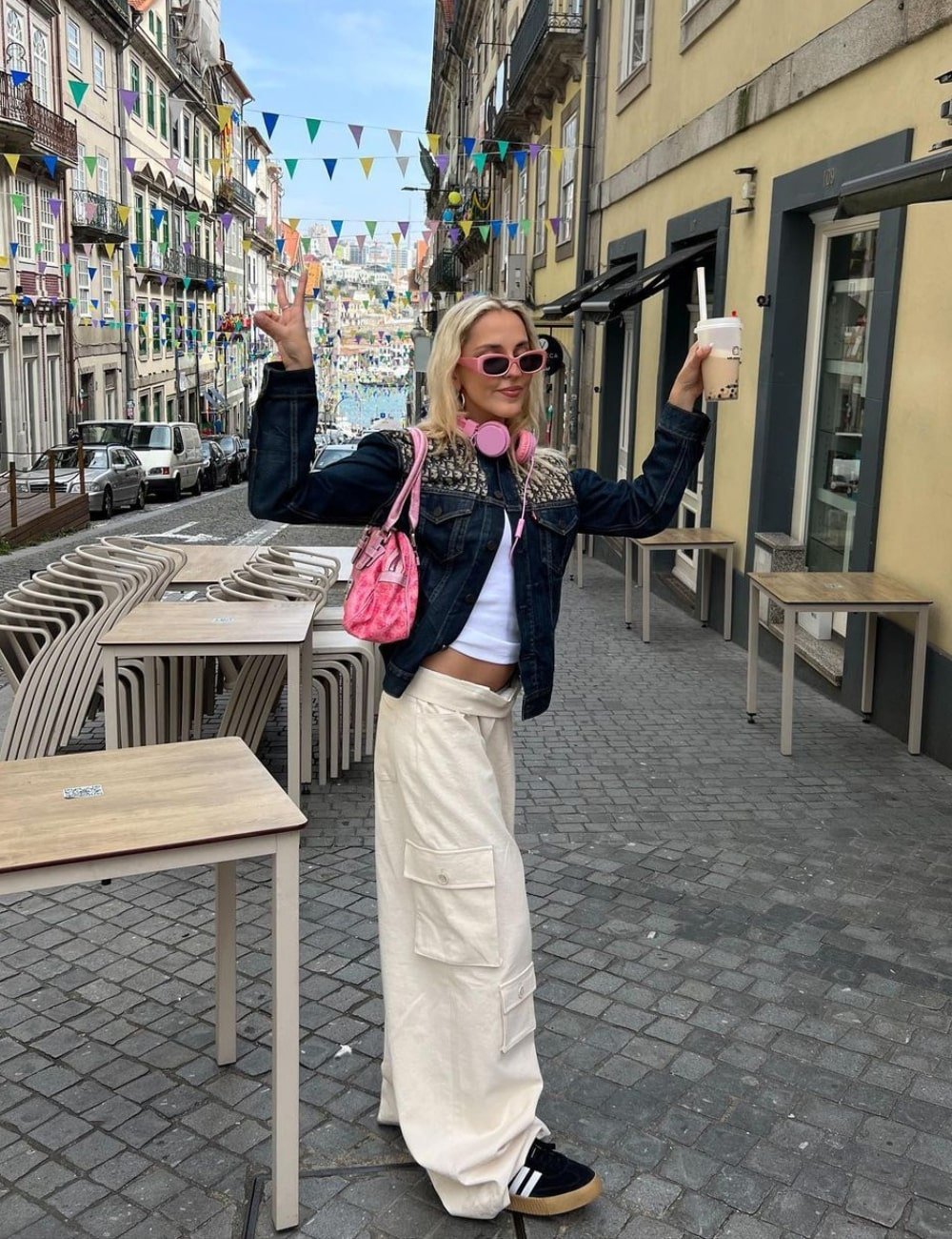 Pessoa posando em rua, vestindo calça cargo larga bege, cropped branco, jaqueta jeans escura e tênis preto Adidas. Acessórios incluem óculos de sol cor-de-rosa, fones de ouvido rosa e bolsa cor-de-rosa. Estilo casual com tendências despojadas e urbanas.