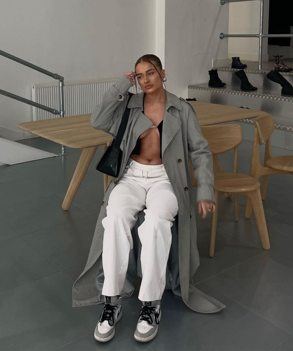 Harriet Slamin - calça branca, pin top preto, tênis e trench coat cinza - trench coat - Outono - sentada em uma cadeira - https://stealthelook.com.br