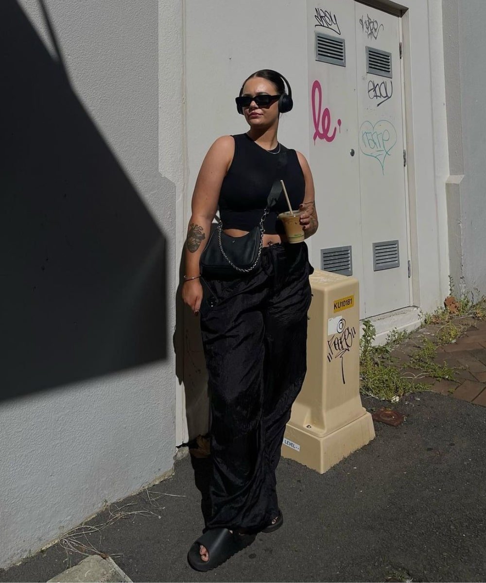 Chelsea Butcher - calça preta, cropped preto, tamanco e óculos escuros - looks novos - Verão - em pé na rua com um fone de ouvido - https://stealthelook.com.br