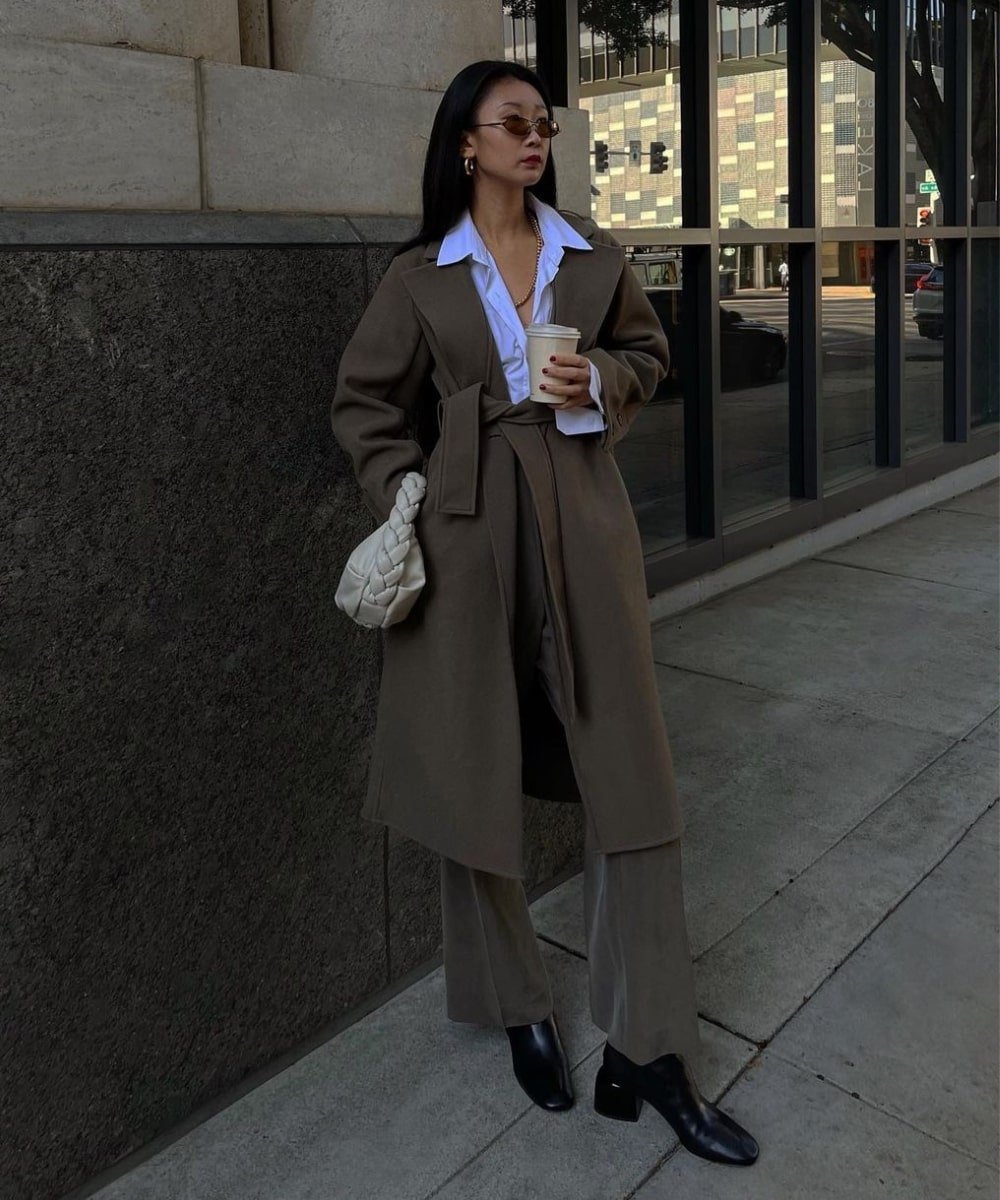 Caroline Lin - calça, camisa branca, sobretudo marrom e botas - como usar cinto - Inverno  - em pé na rua segurando um copo de café na mão esquerda - https://stealthelook.com.br