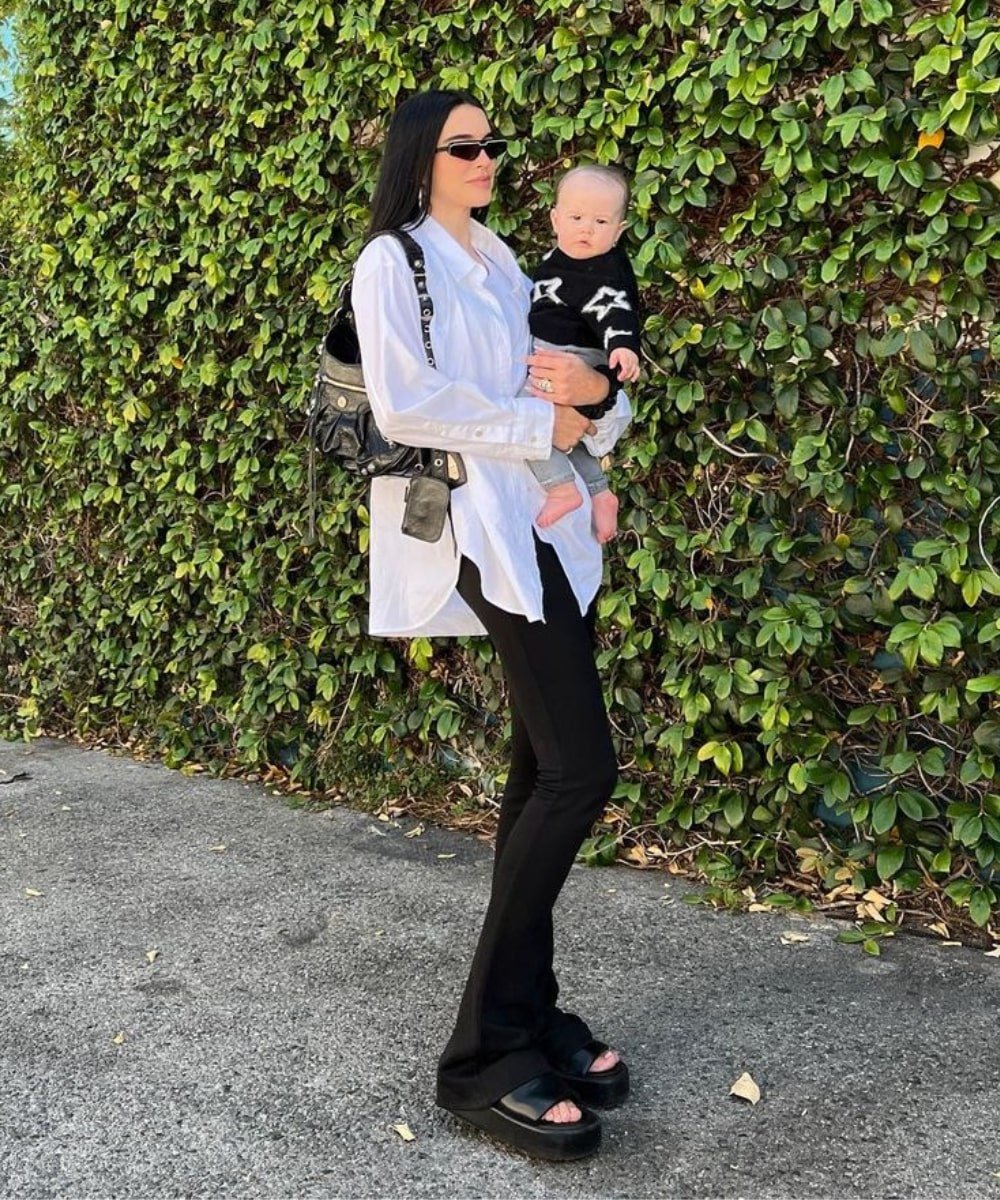 Brittany Xavier - calça preta de yoga, camisa branca oversized e bolsa - calça tendência - Verão - em pé na rua segurando um bebê - https://stealthelook.com.br