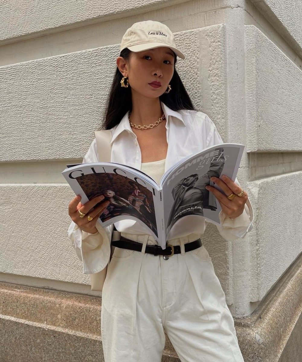 Caroline Lin - calça branca, camisa, cinto preto, bijoux douradas e bone - acessórios de moda - Verão - em pé na rua lendo um jornal aberto - https://stealthelook.com.br