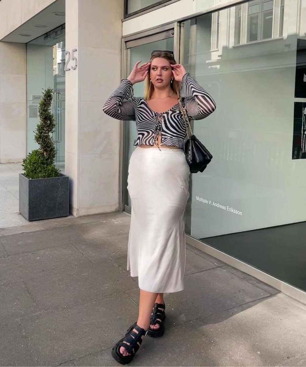 Madison | @Madisoneley - saia midi acetinada, top transparete de animal print e sandálias pretas - looks de verão - Verão - em pé na rua usando óculos de sol - https://stealthelook.com.br