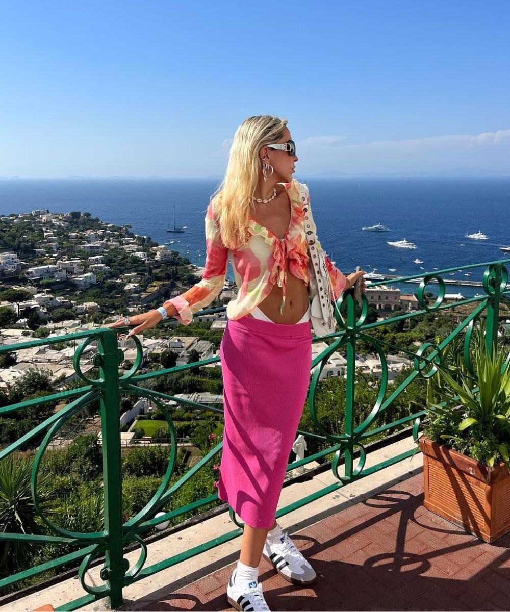 Sofia Coelho - saia midi pink, tênis adidas velosamba e blusa trasnparente estampada - verão 2023 - Verão - em pé em uma varanda com o mar de fundo - https://stealthelook.com.br