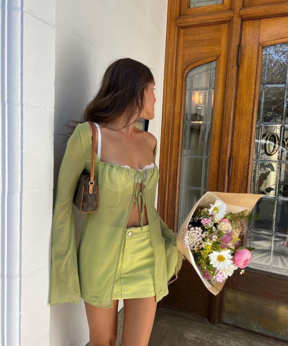 Emma Rose - minissaia verde, bata de amarração transparente e lingerie branca - verão 2023 - Verão - em pé na rua segurando um buque de flores - https://stealthelook.com.br