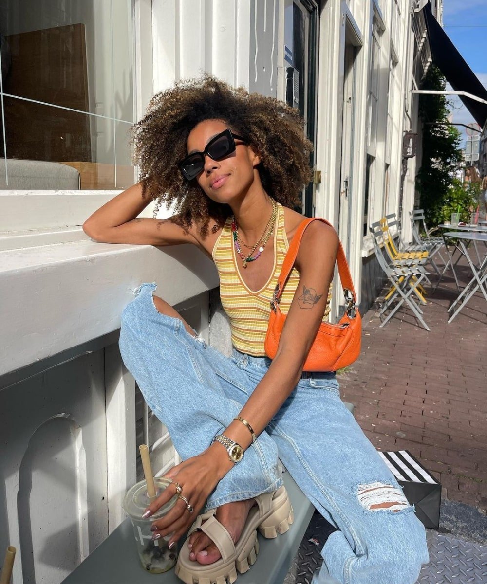 Amaka Hamelijnck - calça jeans, cropped listrado colorido e papetes - como usar calça jeans - Verão - sentada em um banco usando óculos de sol - https://stealthelook.com.br