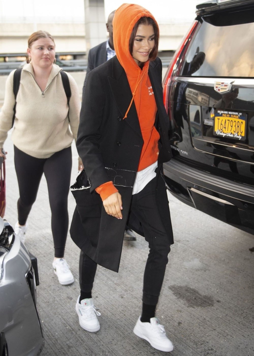 Zendaya - calça preta, tênis, moletom laranja e blazer - look de aeroporto - Inverno  - andando na rua - https://stealthelook.com.br