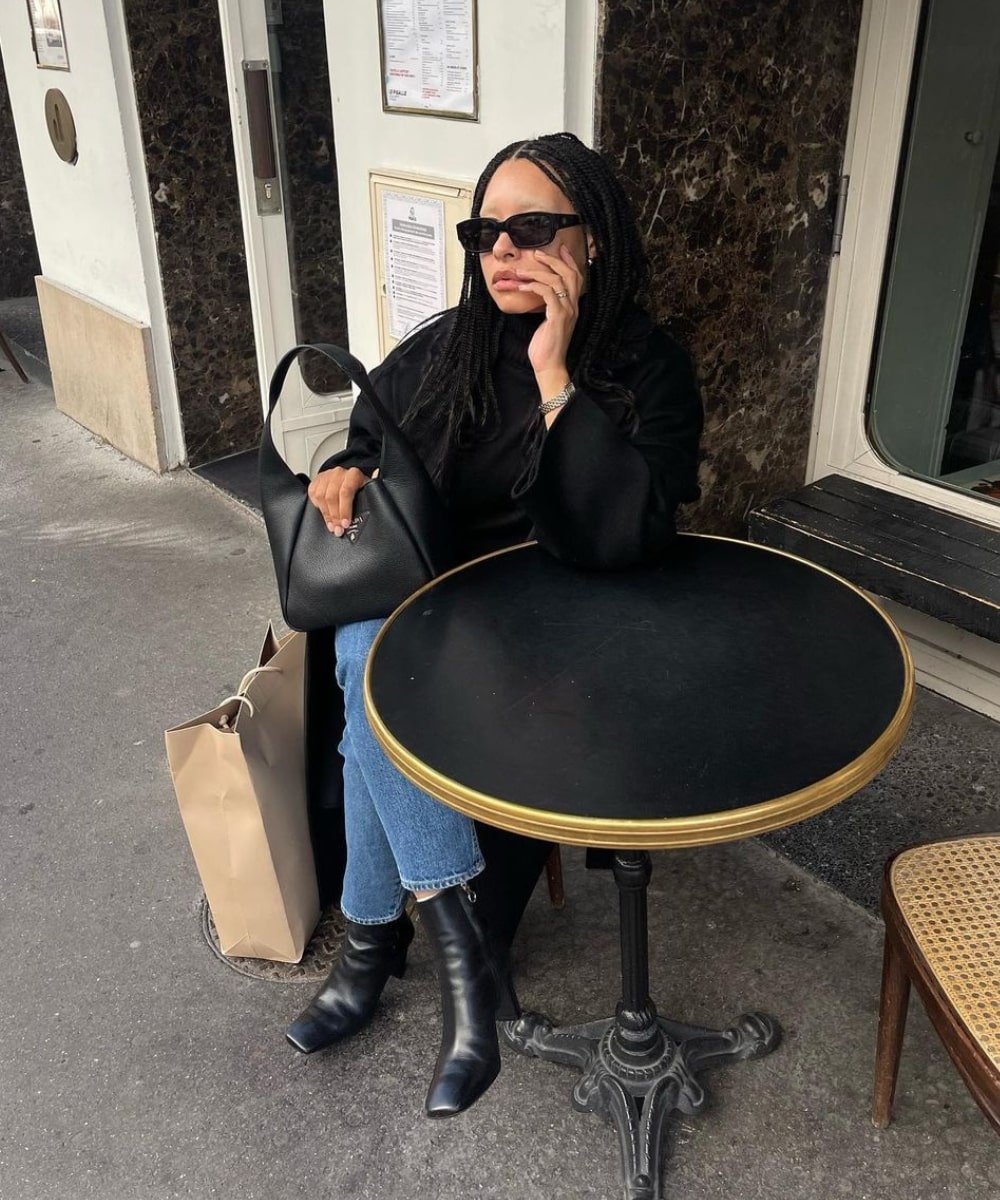 Tanice Elizabeth - calça jeans, bota preta e sobretudo preto - looks com jeans - Inverno  - sentada em um café usando óculos de sol - https://stealthelook.com.br