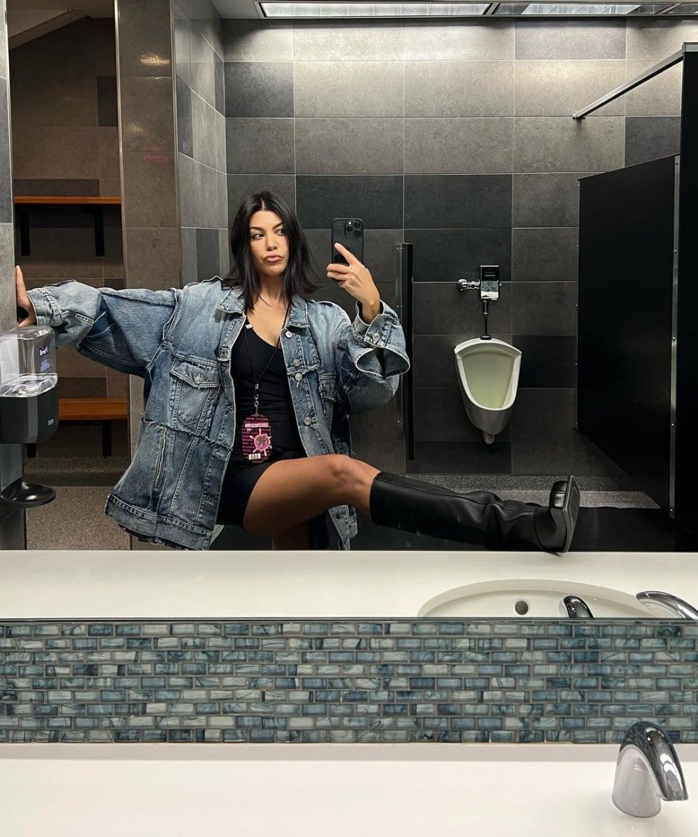 Kourtney Kardashian - jaqueta oversized jeans, vestido curto preto e botas de cano longo - Kourtney Kardashian - Outono - foto em um banheiro com a perna em cima da pia - https://stealthelook.com.br