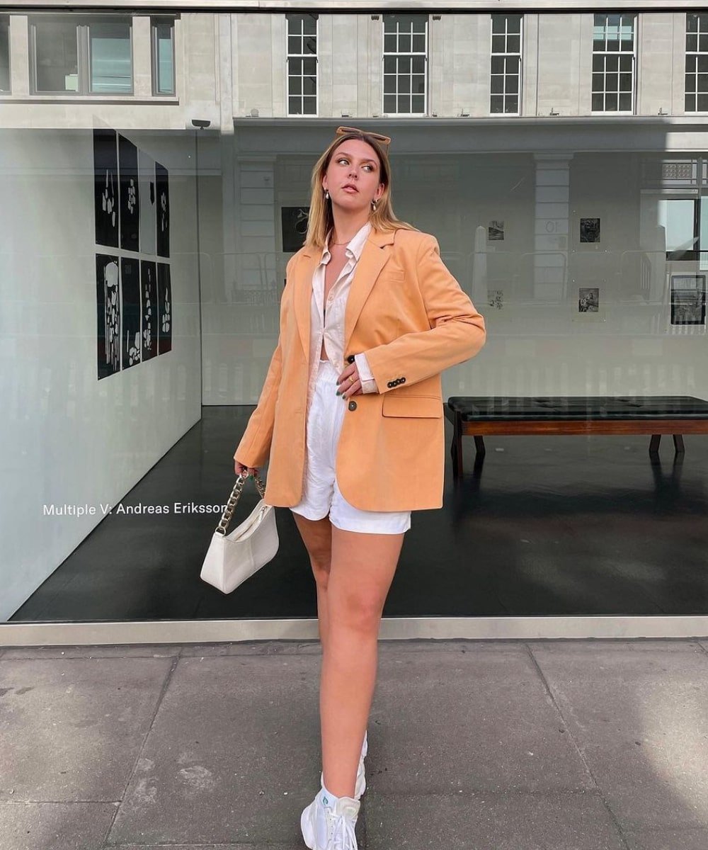 Madison | @Madisoneley - short branco, camisa e blazer laranja - peças indispensáveis - Verão - em pé na rua segurando uma bolsa branca - https://stealthelook.com.br