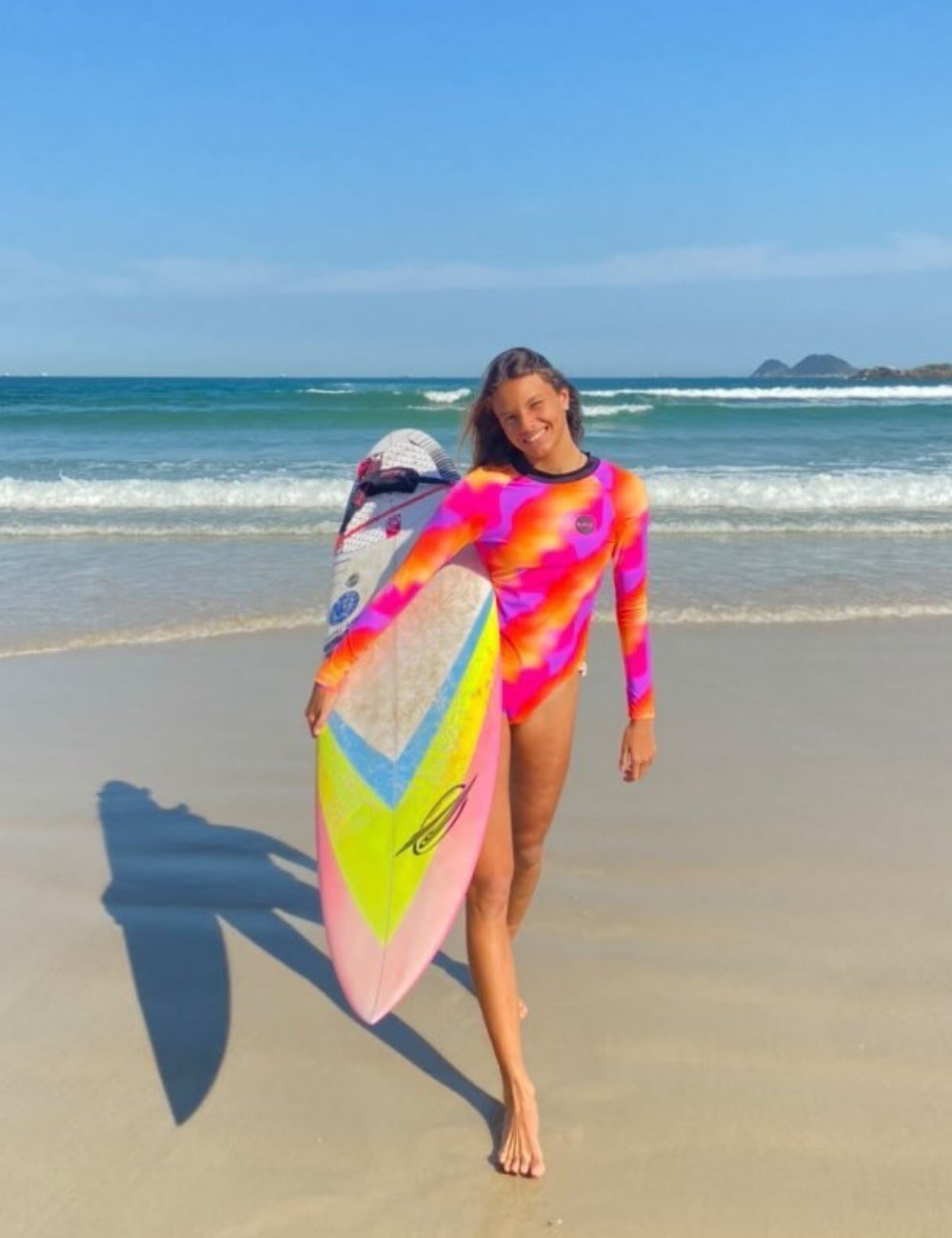 Somos 1 - maiô de manga longa estampado e colorido - tendências de verão - Verão - mulher andando na praia com uma prancha de surfe na mão - https://stealthelook.com.br