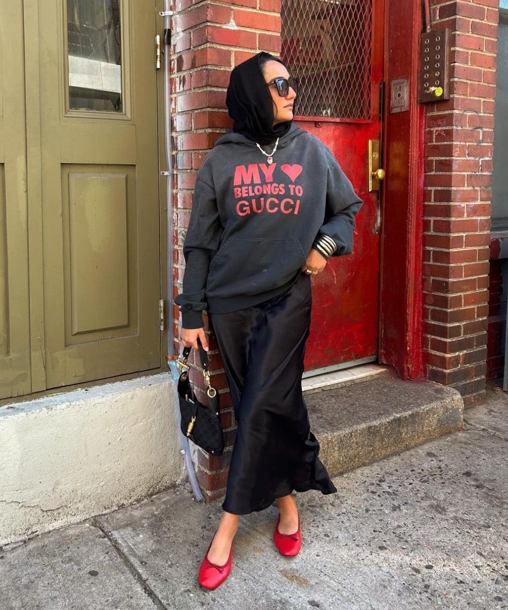 Yusra Siddiqui - saia longa preta, moletom com escrito, sapatilhas vermelhas e óculos escuros - anos 80 - inverno - em pé na rua olhando para o lado - https://stealthelook.com.br