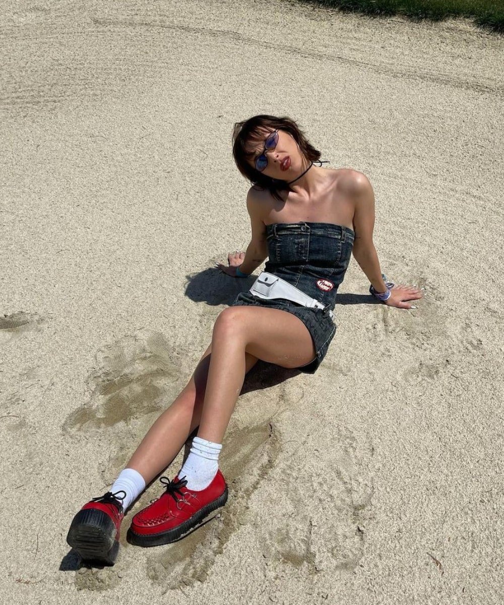 Emma Chamberlain - conjunto de minissaia e cropped jeans, sapato vermelho e óculos escuros - looks do Coachella - verão - sentada na areia com as mãos apoiadas para trás - https://stealthelook.com.br