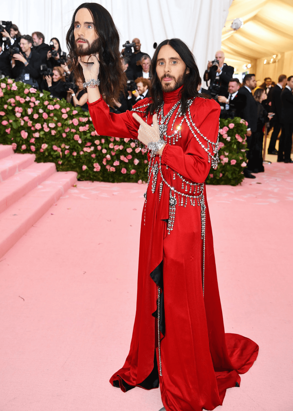 Jared Leto - vestido vermelho longo e calça roxa metalizada - Met Gala - primavera - homem segurando uma replica de sua cabeça com a mão no alto - https://stealthelook.com.br