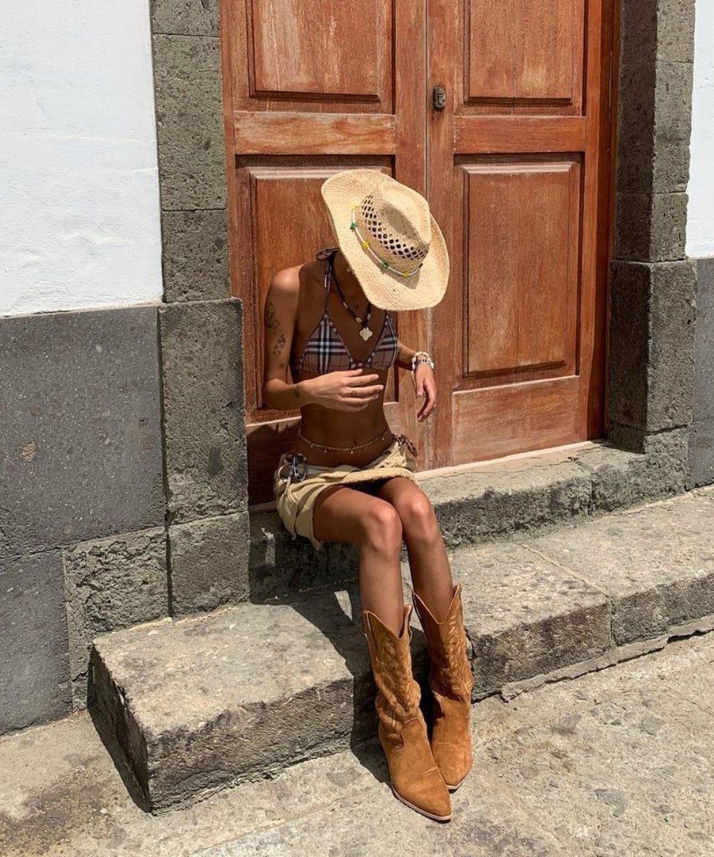 Daisy Birchall - minissaia, biquini e botas cowboy - tendências do TikTok - verão - sentada em uma escada na rua - https://stealthelook.com.br