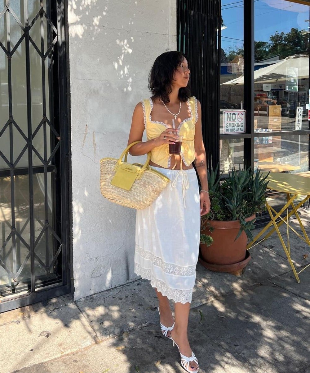 Stephanie Arant - top amarelo, saia midi off white e bolsa de palha - tendências do TikTok - verão - em pé na rua segurando uma bolsa - https://stealthelook.com.br