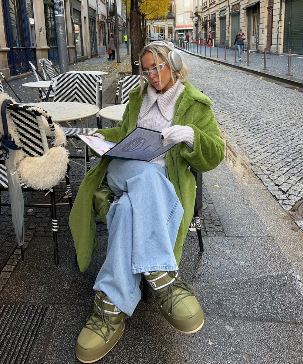 Sofia Coelho - calça jeans, moon boots, casaco bege de cola e casaco longo verde - moon boots - inverno - sentada em uma cadeira na rua com um cardápio na mão - https://stealthelook.com.br