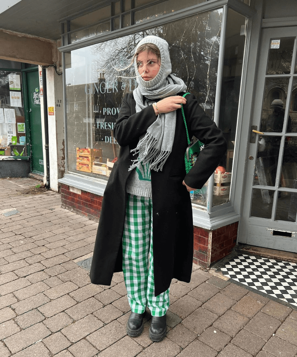 Abigail Liddiard - calça xadrez, tricot, sobretudo preto, balaclava cinza e bota preta tratorada - looks com xadrez - inverno - em pé na rua - https://stealthelook.com.br