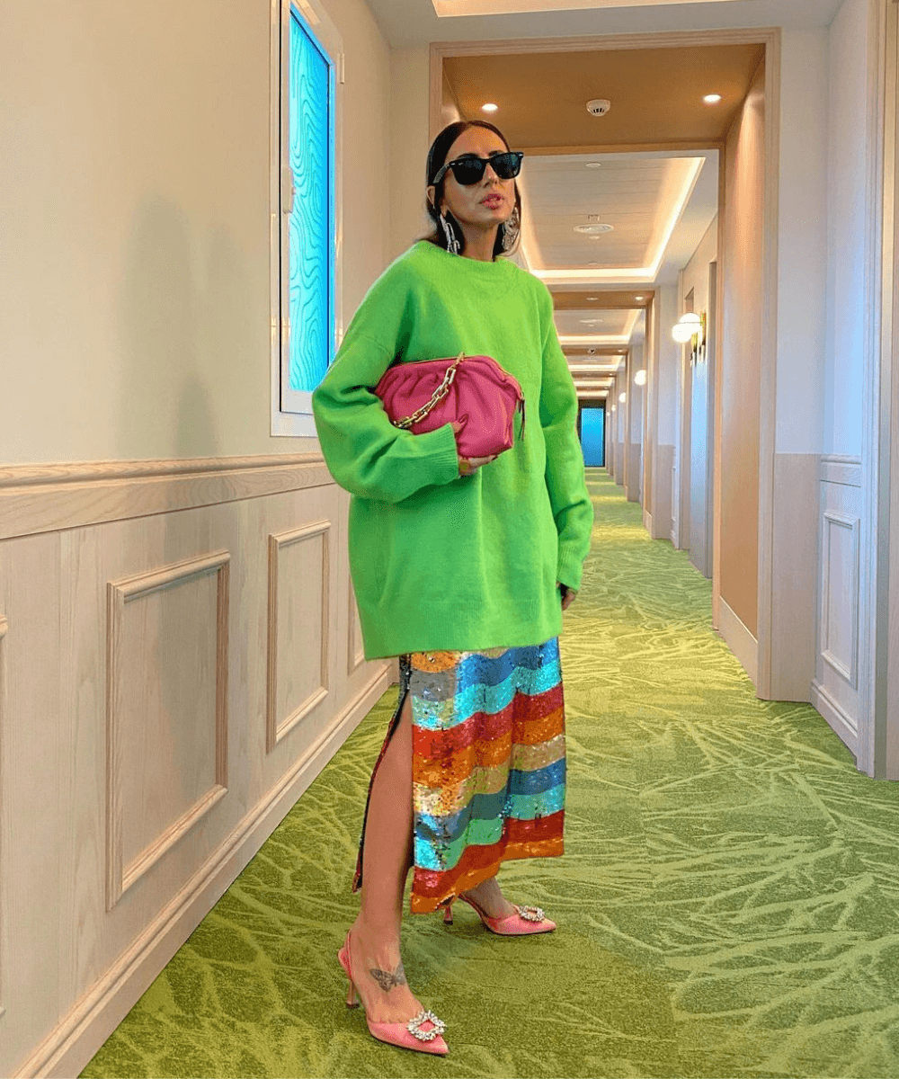 Laura Eguizabal - tricot verde, saia de paete colorida e óculos de sol - looks de festa - inverno - em pé em um corredor - https://stealthelook.com.br