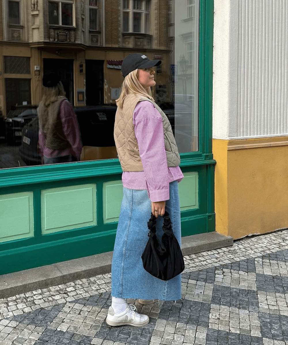 @lillibethchen - saia midi jeans, camisa listrada rosa, colete bege, boné e tênis adidas - saia midi - inverno - mulher loira de lado andando na rua - https://stealthelook.com.br