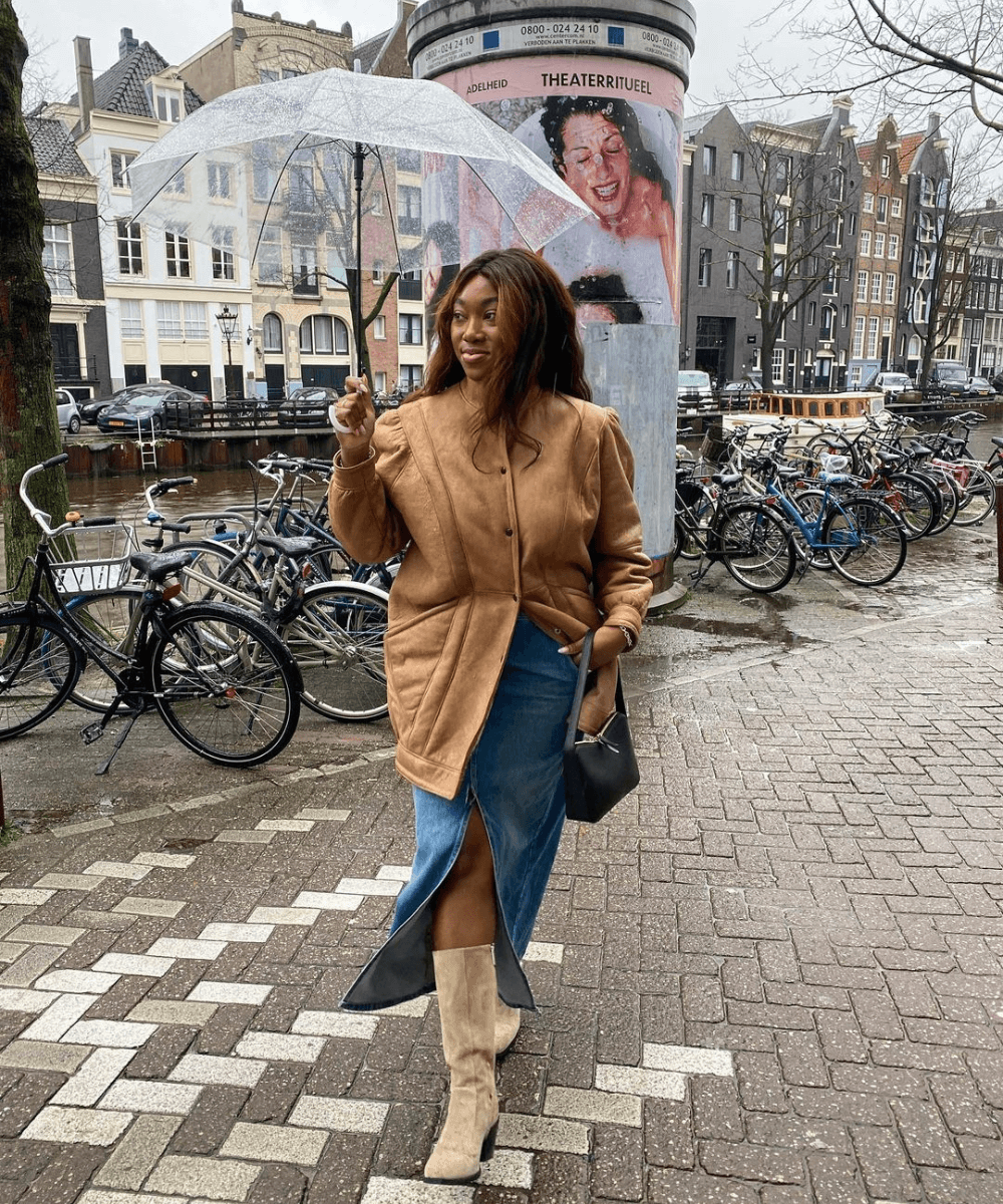 Nnenna Echem - saia jeans, bota bege e jaqueta marrom - looks de festa junina - inverno - mulher andando na rua segurando um guarda-chuva transparente - https://stealthelook.com.br