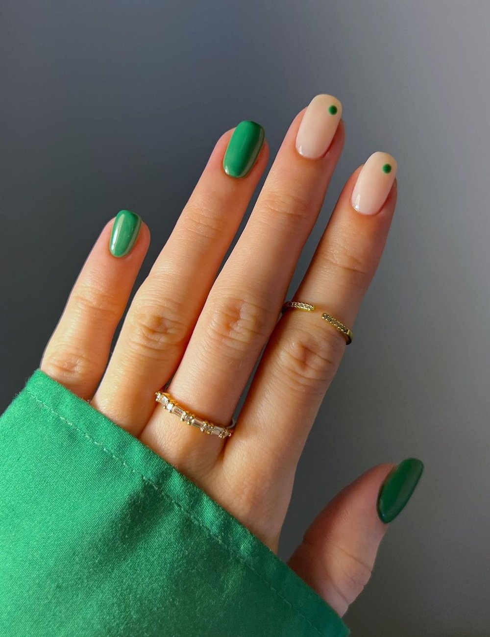 Unhas com nail art verde e nude com ponto verde, um mix moderno e minimalista. Mão vestindo camisa verde mostra acessórios elegantes como anéis delicados dourados, destacando-se como tendência de moda em esmaltação e acessórios.