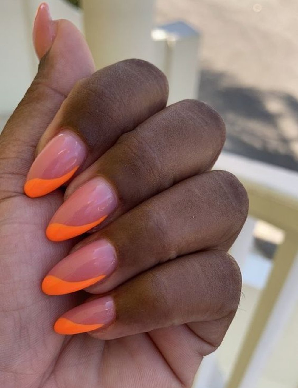 Nail art com esmalte nude e pontas decoradas em laranja fluorescente. O design apresenta uma abordagem moderna e vibrante para a clássica manicure francesa, seguindo tendências de moda que valorizam cores chamativas e contrastes nas unhas.