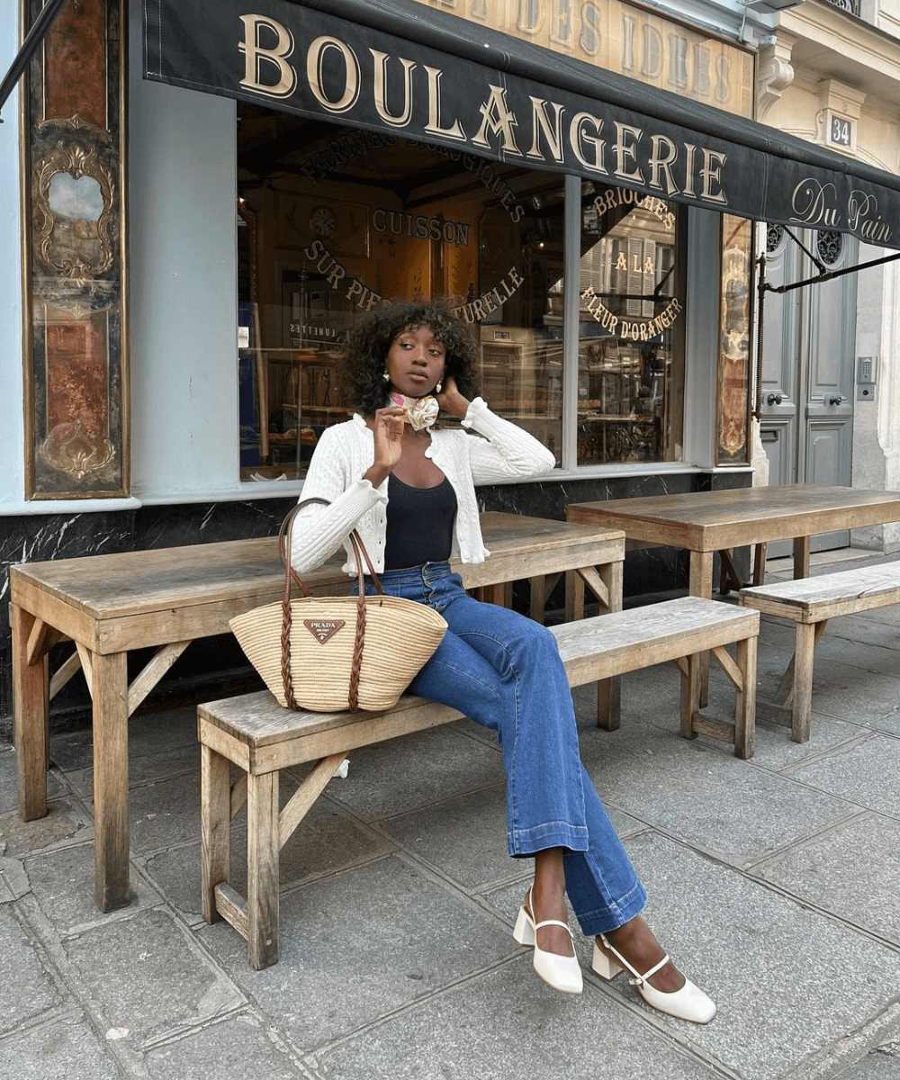Emmanuelle Koffi - calça jeans, regata preta, cardigã branco e sandália mary jane branca - sapato tendência - outono - mulher negra sentada em um banco na rua olhando para o lado - https://stealthelook.com.br