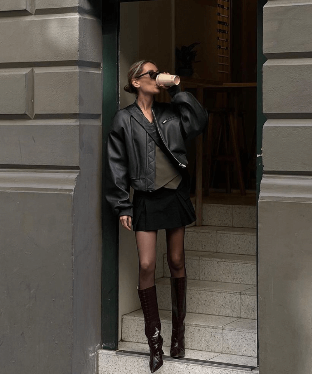 Jess Alizzi - saia curta, bota de cano longo e jaqueta de couro - jaqueta biker - inverno - mulher em pé na rua bebendo café - https://stealthelook.com.br