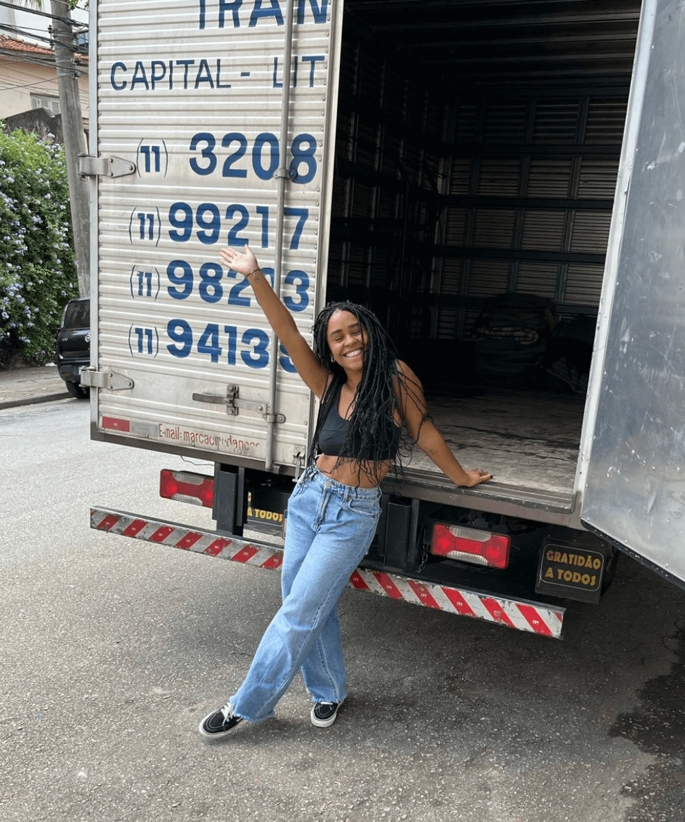 Inaê Ribeiro - calça jeans e camiseta preta - mudar para São Paulo - verão - mulher negra de tranças em pé na rua apoiada em um caminhão de mudança sorrindo - https://stealthelook.com.br