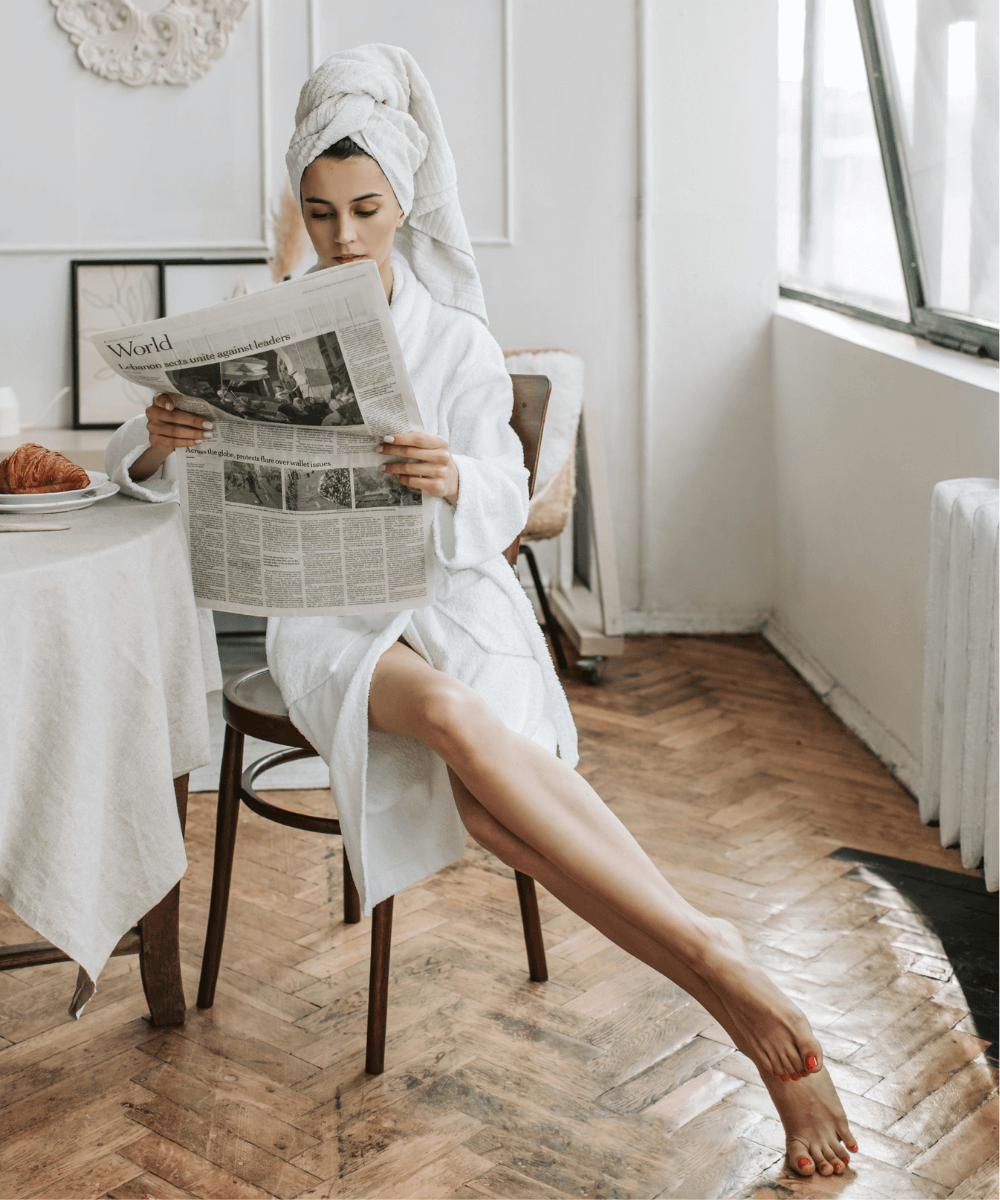 Vlada Karpovich - roupão branco - itens de inverno - inverno - mulher de roupão branco e toalha na cabeça, sentada em uma cadeira enquanto lê um jornal - https://stealthelook.com.br
