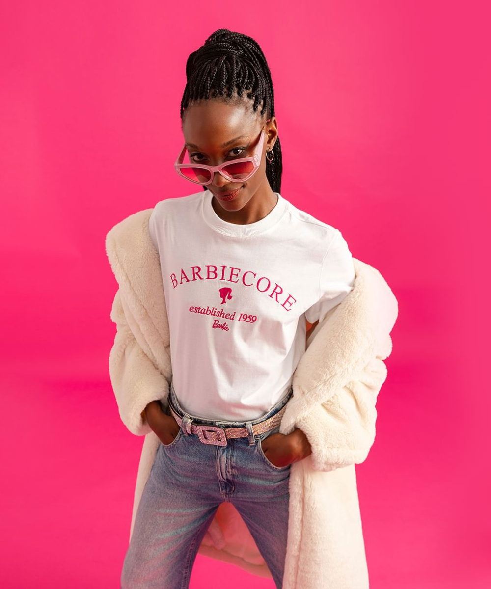 Coleção-cápsula C&A e Barbie - t-shirt branca com escrito em rosa, jaqueta rosa e calça jeans - sapato da Barbie - inverno - mulher negra andando na rua usando óculos de sol em um fundo rosa - https://stealthelook.com.br