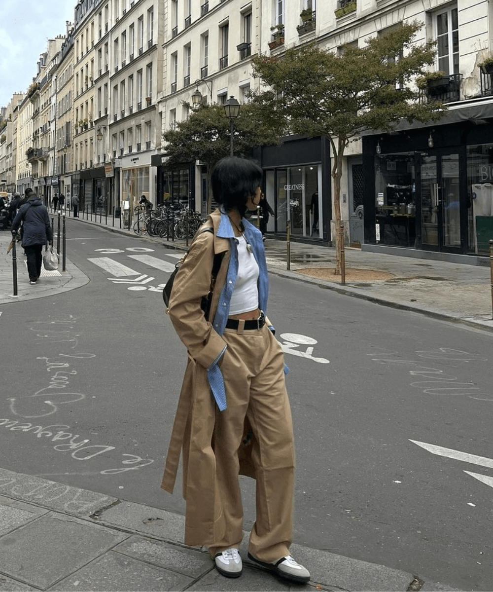 Pessoa com cabelo curto preto e máscara caminha em uma rua. Veste calça wide leg bege, trench coat bege sobre camisa azul e top branco. Complementa o visual fashionista com cinto preto, tênis branco, mochila preta e estilo urbano despojado.