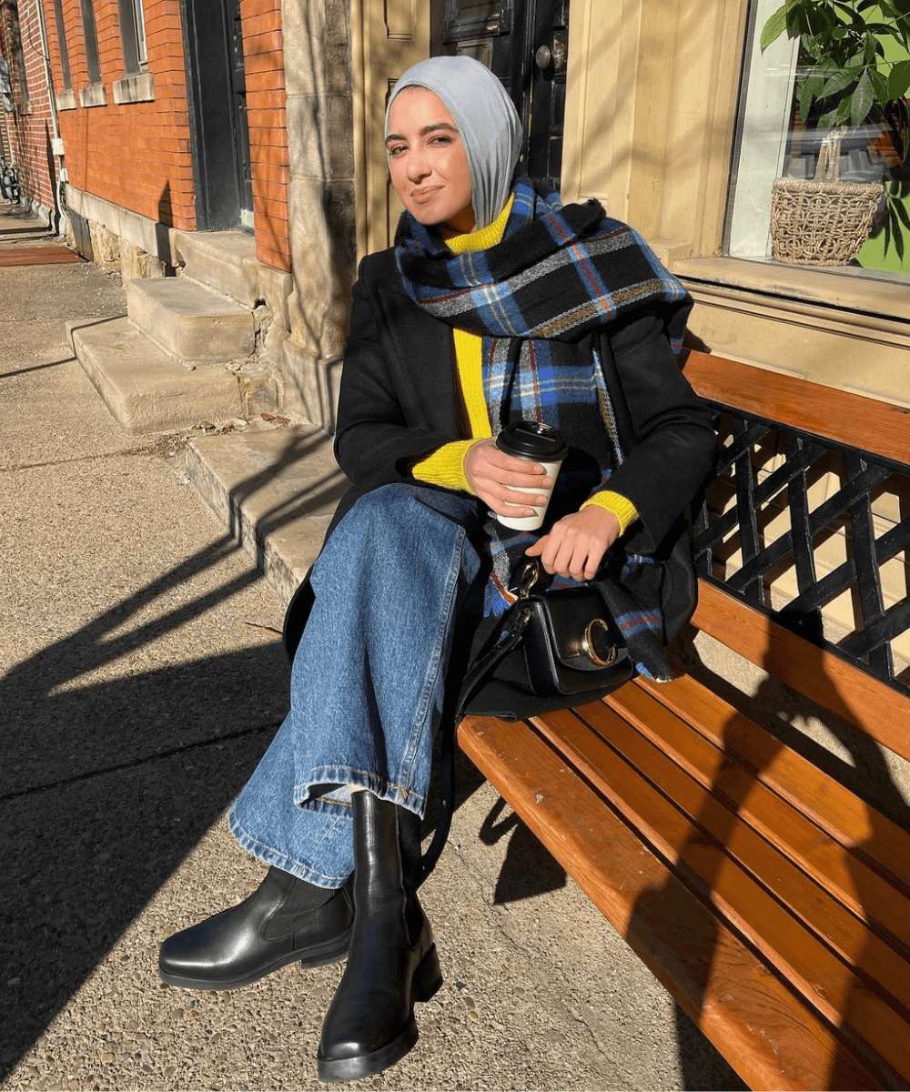 @summeralbarcha - calça jeans, bota preta, tricot amarelo e sobretudo preto - looks com tricô - inverno - mulher sentada em um banco na rua tomando café - https://stealthelook.com.br
