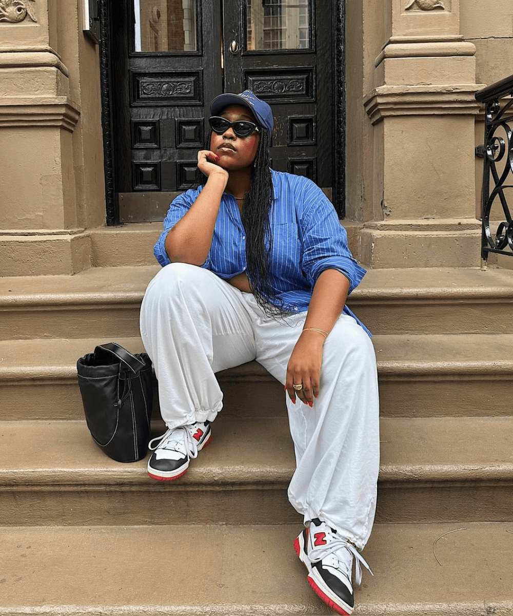 Aniyah Morinia - calça branca, tênis e camisa azul listrada - looks fáceis - outono - mulher negra sentada em uma escada usando boné e óculos de sol - https://stealthelook.com.br
