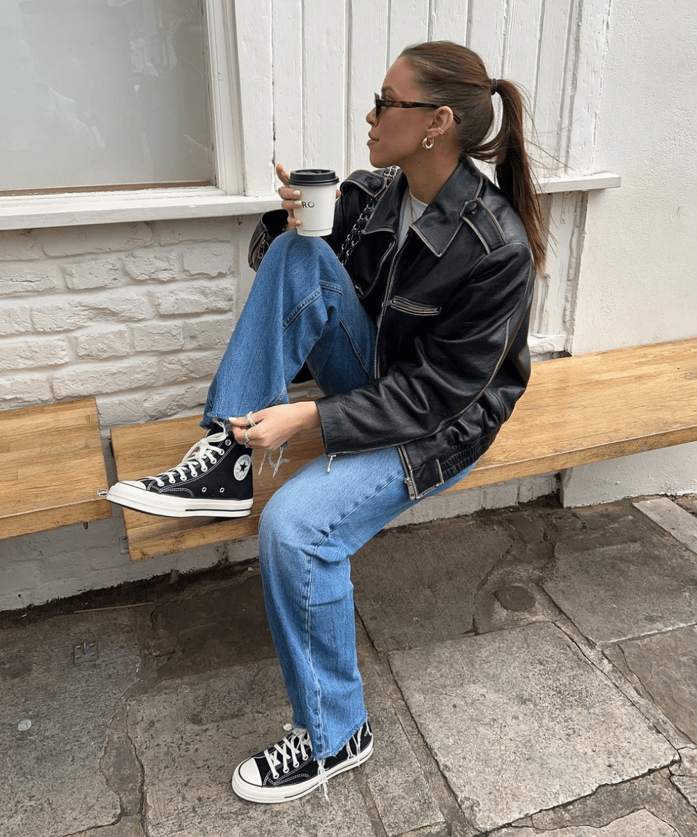 Jamie Firth - calça jeans, tênis converse preto, blusa branca e jaqueta preta de couro - tênis da moda - inverno - mulher sentada em um banco na rua com uma perna levantada e segurando um copo de bebida transparente - https://stealthelook.com.br