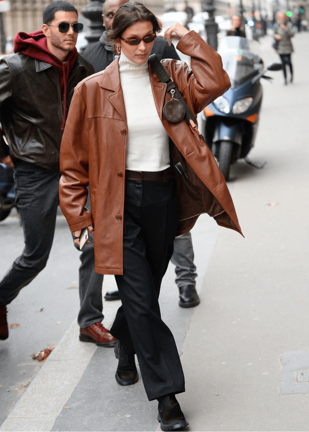 Bella Hadid - calça preta, blusa branca de manga longa, blazer de couro marrom e botas pretas - Bella Hadid - inverno - modelo andando na rua com uma bolsa no ombro e usando óculos escuros - https://stealthelook.com.br