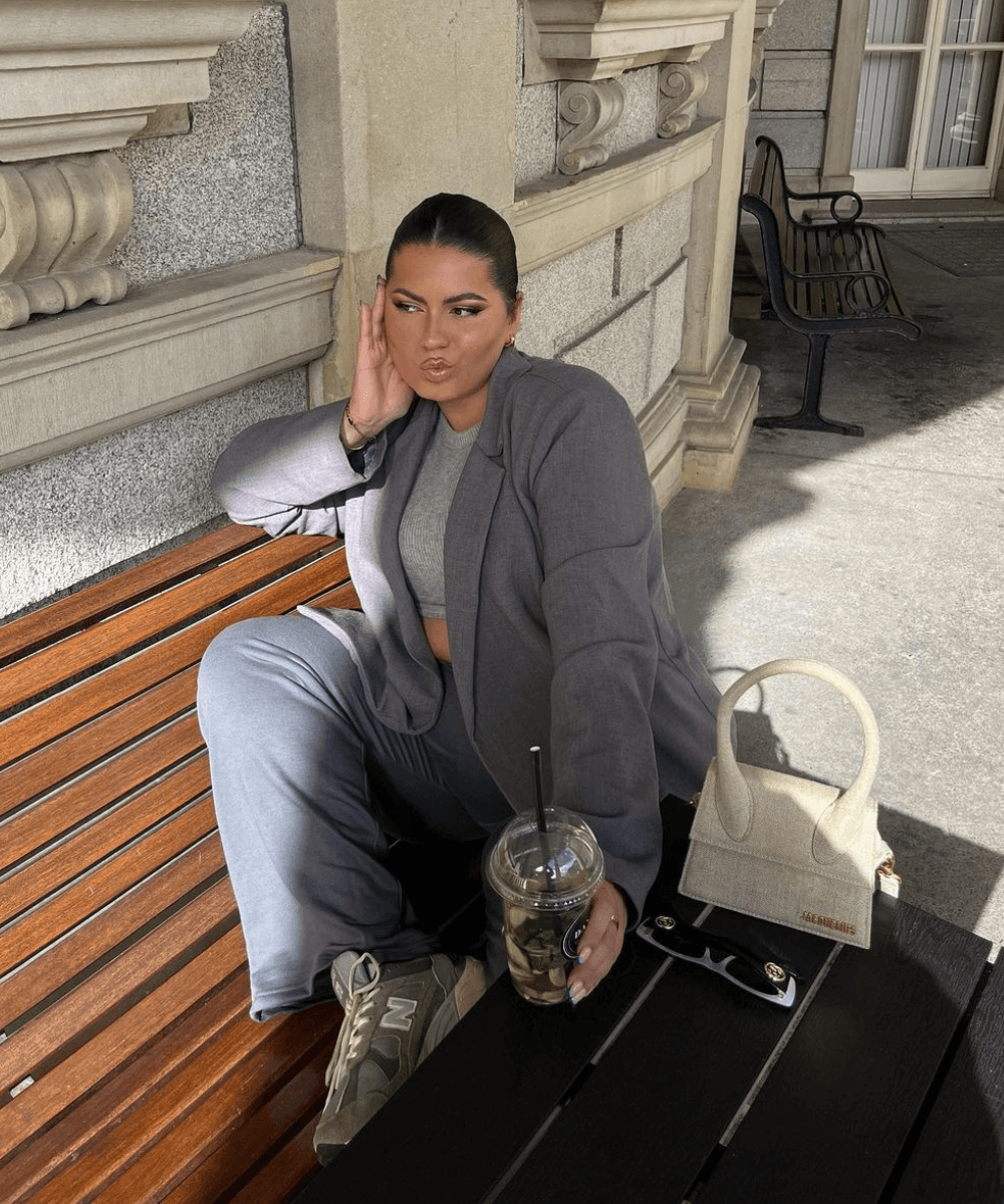 Freya Broni - conjunto cinza de alfaiataria, cropped e tênis - look de trabalho - outono - mulher sentada em um banco na rua com uma perna levantada e segurando um copo de bebida transparente - https://stealthelook.com.br