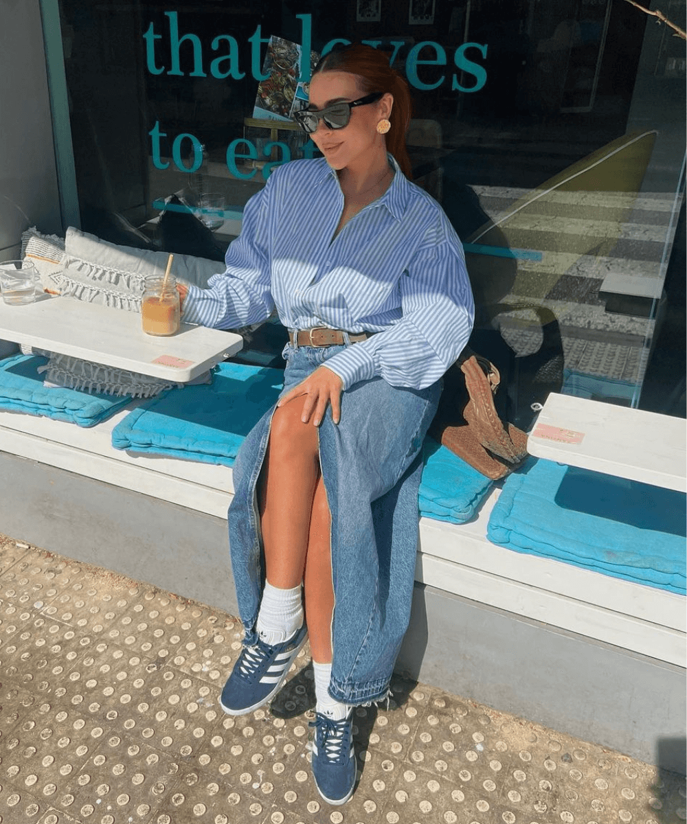 Ellie Beatrice Joslin - saia longa jeans, camisa listrada azul e tênis adidas - tendência de moda - outono - mulher sentada em um banco na rua com as pernas cruzadas usando óculos de sol - https://stealthelook.com.br