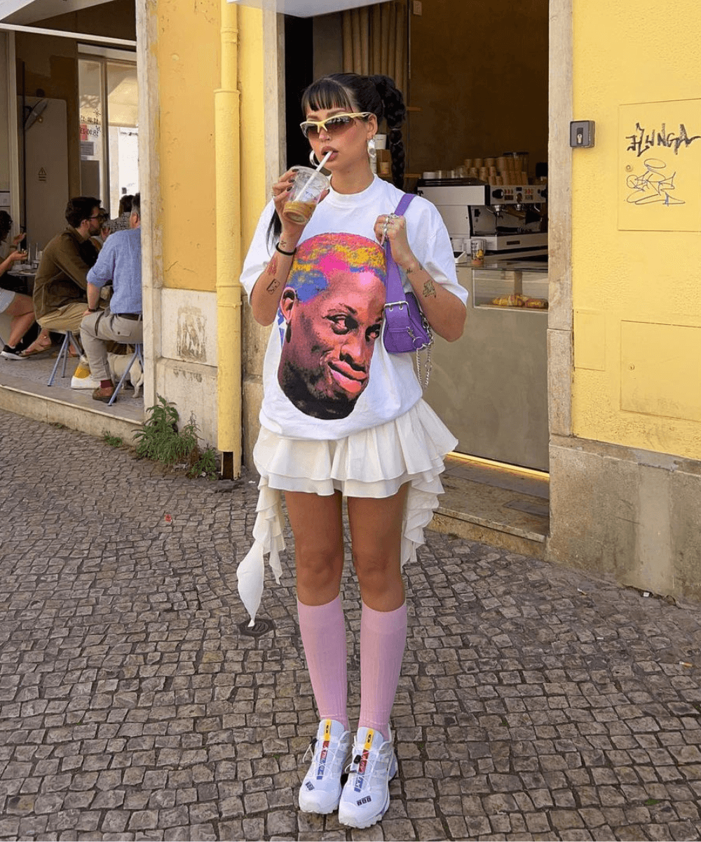 Ines Silva | @irisloveunicorns - tshirt estampada, saia de babados e meia longa rosa com tênis branco - tendência Y2K - verão - mulher morena em pé na rua bebendo em um copo com canudo - https://stealthelook.com.br