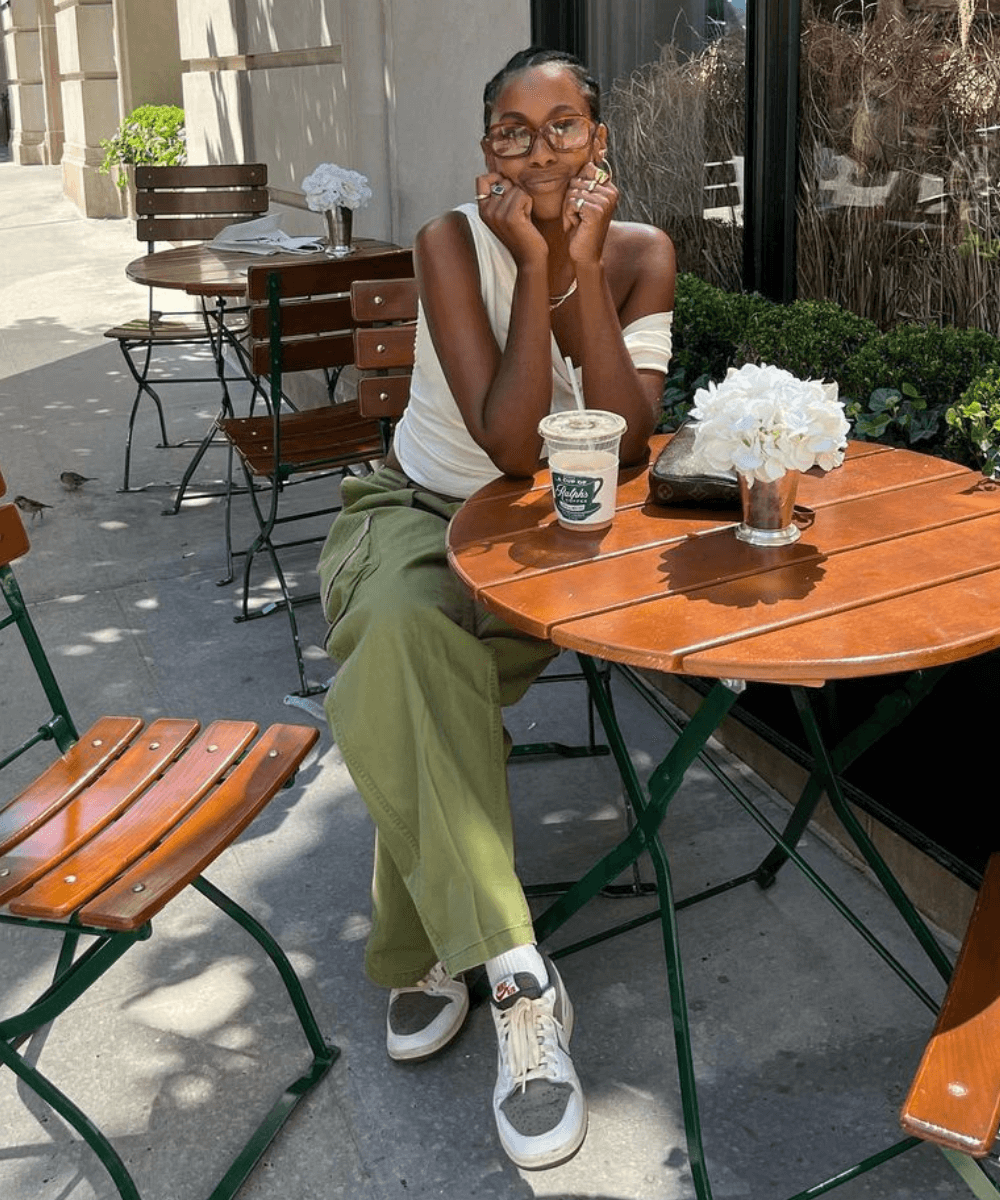 Claire Most - calça verde cargo, tênis, blusa drapeada e óculos de sol - anos 2000 - verão - mulher negra sentada em um café na rua usando óculos de sol - https://stealthelook.com.br