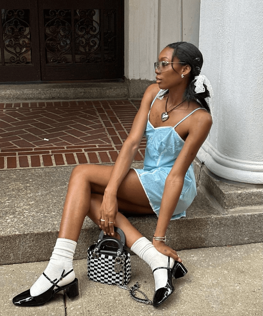Chanel Mckinsie - vestido azul curto, meias aparentes, sandália slinback preta e óculos escuros - sandálias da moda - verão - mulher negra sentada na sua com as pernas cruzadas com a cabeça virada para o lado - https://stealthelook.com.br