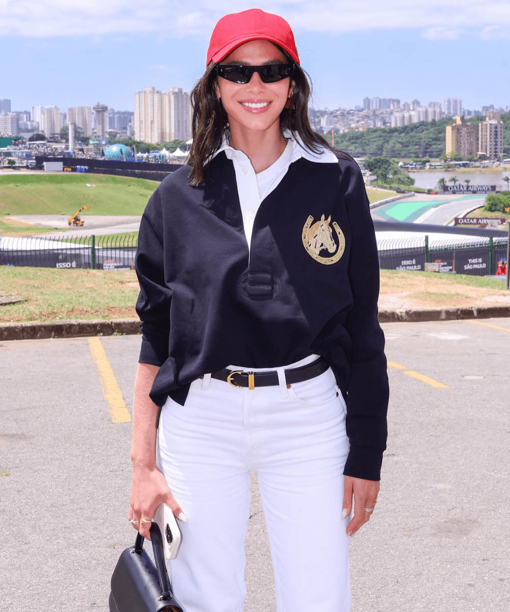 Bruna Marquezine - calça branca, boné vermelho, blusa marinho e bolsa de mão - looks do GP da Fórmula 1 - primavera - mulher de óculos em pé na rua sorrindo - https://stealthelook.com.br