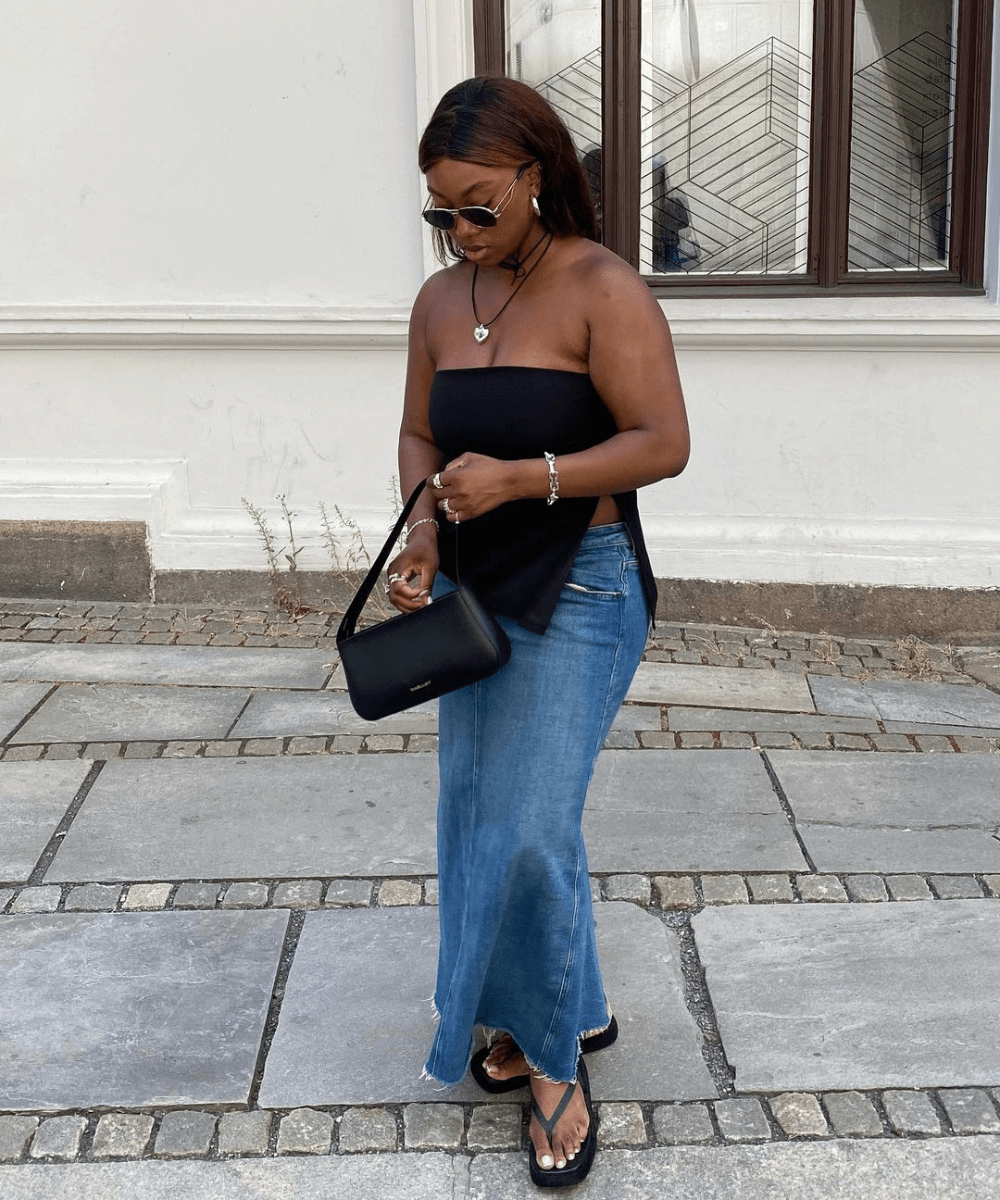 Nnenna Echem - saia midi jeans, top sem alças preto e chinelo preto - tendências polêmicas - verão - mulher negra em pé na rua mexendo na bolsa - https://stealthelook.com.br