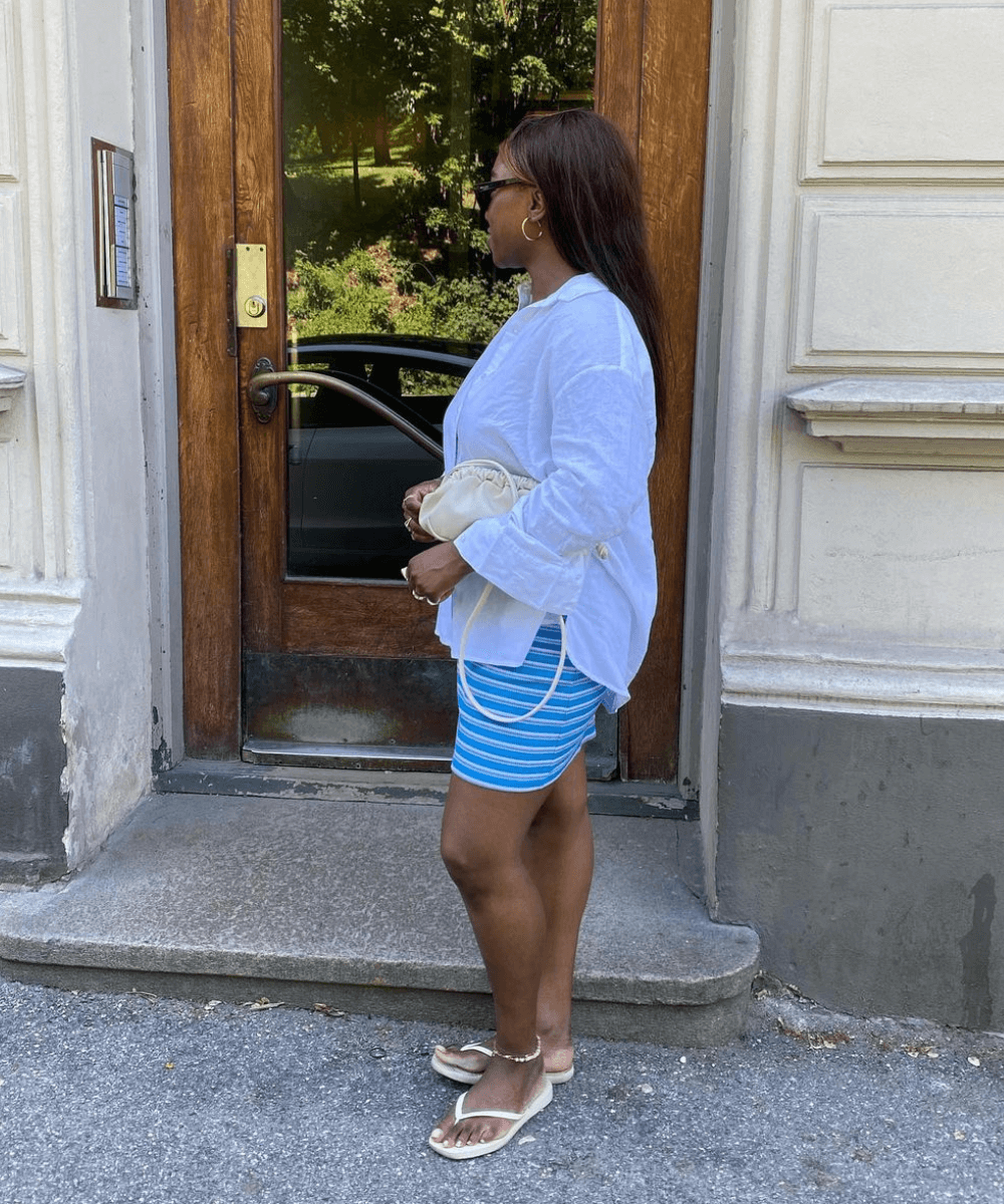 Nnenna Echem - minissaia azul, chinelos e camisa branca oversized - looks com chinelo - verão - mulher negra de lado em pé na rua - https://stealthelook.com.br